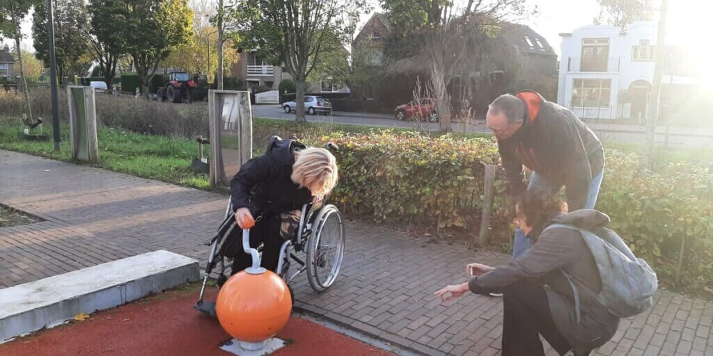 Playground. Person in wheelchair plays with a music ball. Two people comment on the action.
